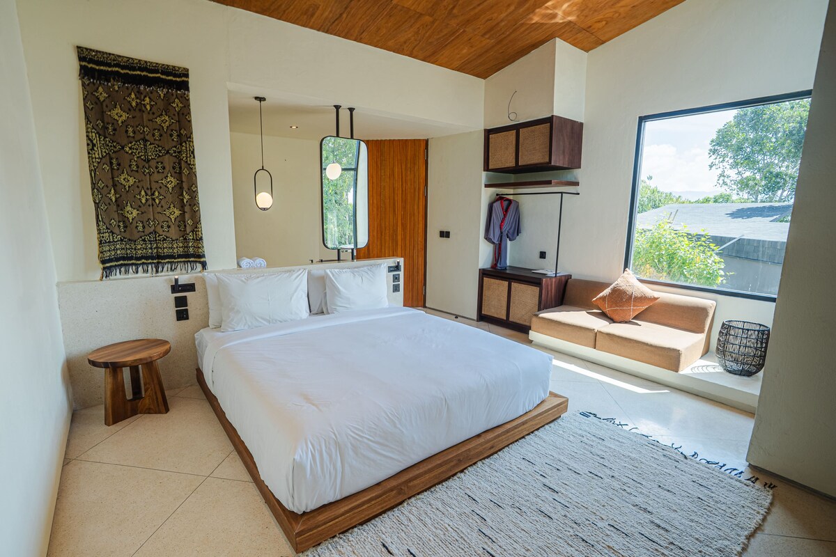 Un dormitorio moderno con una cama grande, un taburete pequeño, un sofá, una lámpara colgante y vistas a través de una ventana.