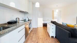 Cuina moderna amb armaris blancs, taulell de granit i forn integrat. A la zona de sala d'estar hi ha un terra de fusta i un sofà gris amb coixins grocs.