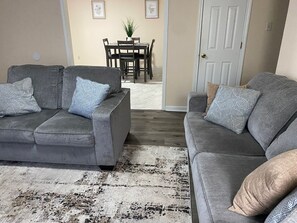 Un salon avec deux canapés gris, un tapis à motifs et un coin repas comprenant une table et des chaises.