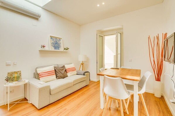 Un salon moderne avec un canapé, une table à manger et des chaises. On y trouve une petite table d’appoint avec une lampe et un cadre photo.