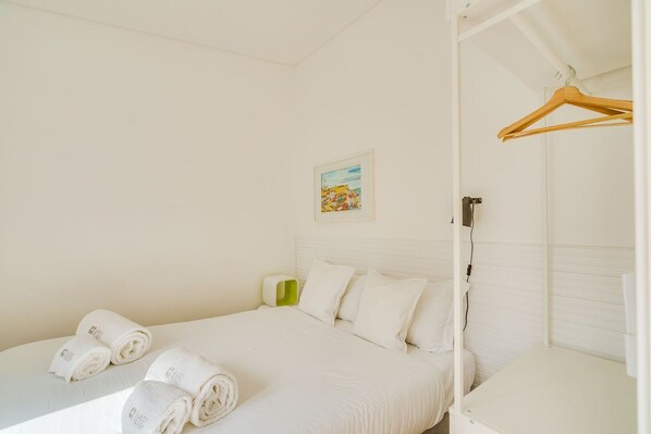 A bedroom with a bed, two rolled towels, a bedside table, a hanging wooden hanger, and a framed picture on the wall.
