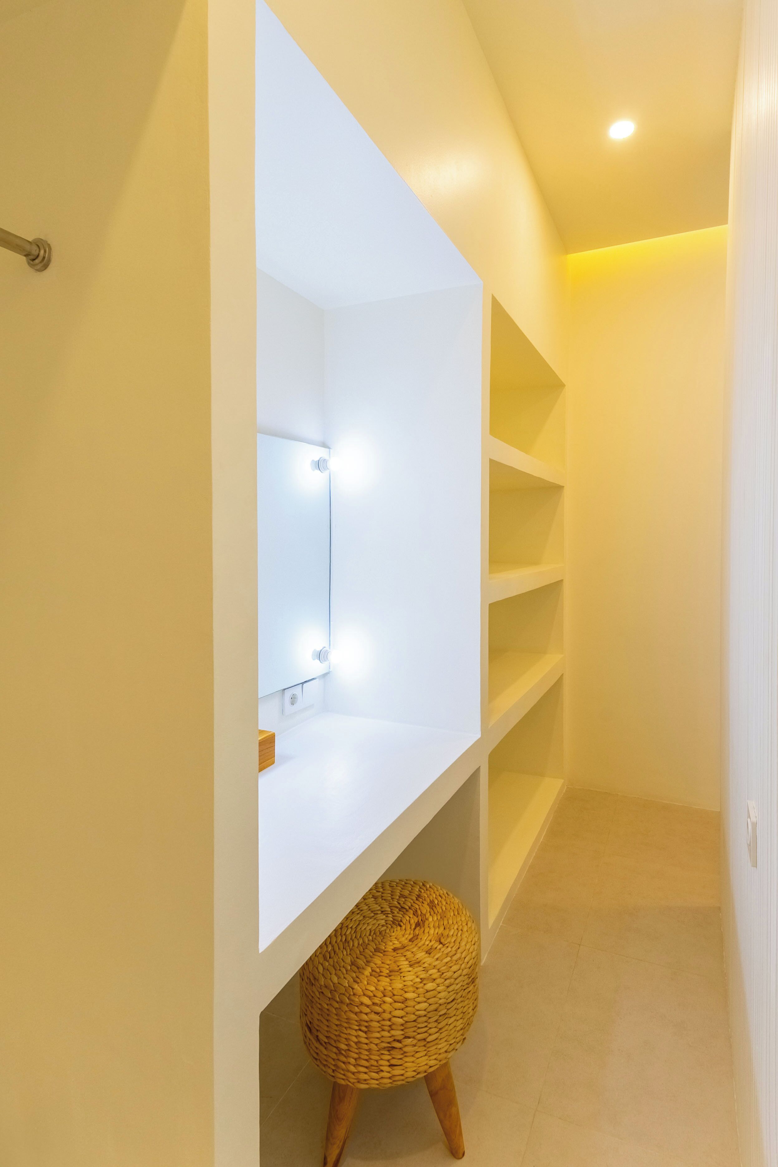 A narrow hallway with a white vanity mirror with lights, a wicker stool, and built-in shelves.