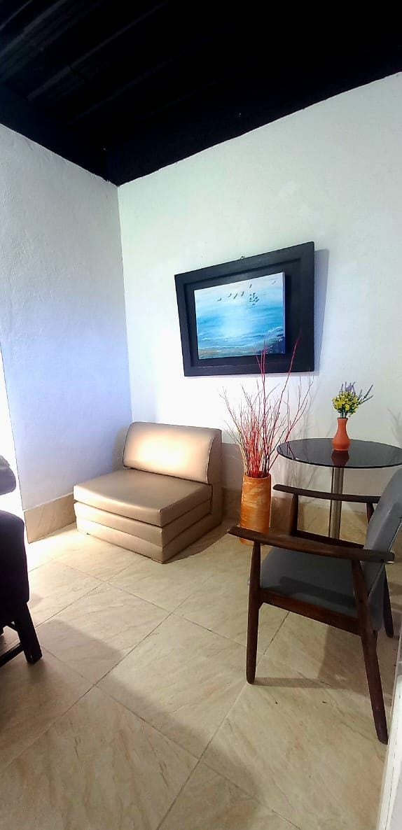 Un salón con un sofá color beige, una mesita redonda con una silla, un televisor montado en la pared y un florero con plantas.