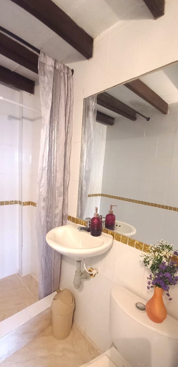 Une salle de bain avec un lavabo blanc, un miroir et des toilettes. On y trouve un rideau de douche, un vase avec des fleurs et une poubelle.