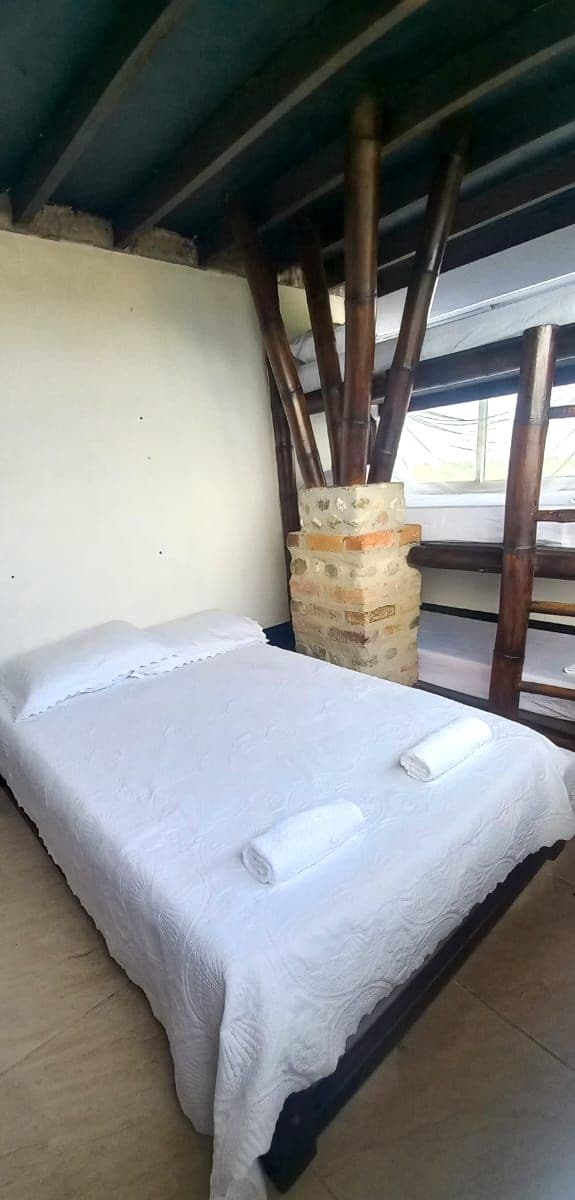 Una cama individual con una colcha blanca y dos toallas enrolladas, un pilar de ladrillo y una estructura de techo de madera.
