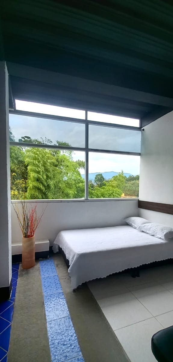 A bedroom with a large window, a bed, a small table, and a vase with plants.