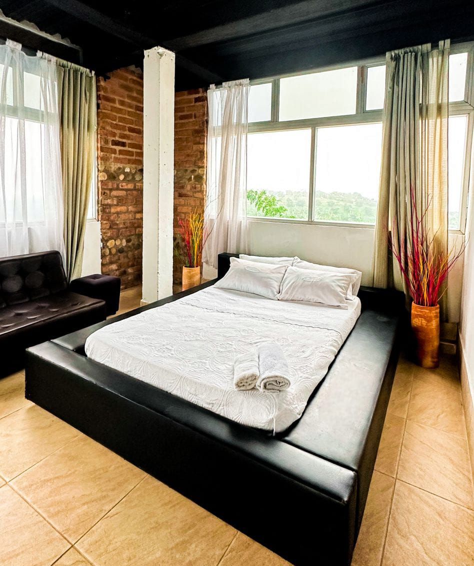 A bedroom with a large bed, a dark sofa, and a view of greenery through the window.
