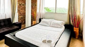 A bedroom with a large bed, a dark sofa, and a view of greenery through the window.