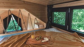 Una habitación acogedora con cama, ventana con vistas a árboles, techo de madera y una mesa de madera con comida.