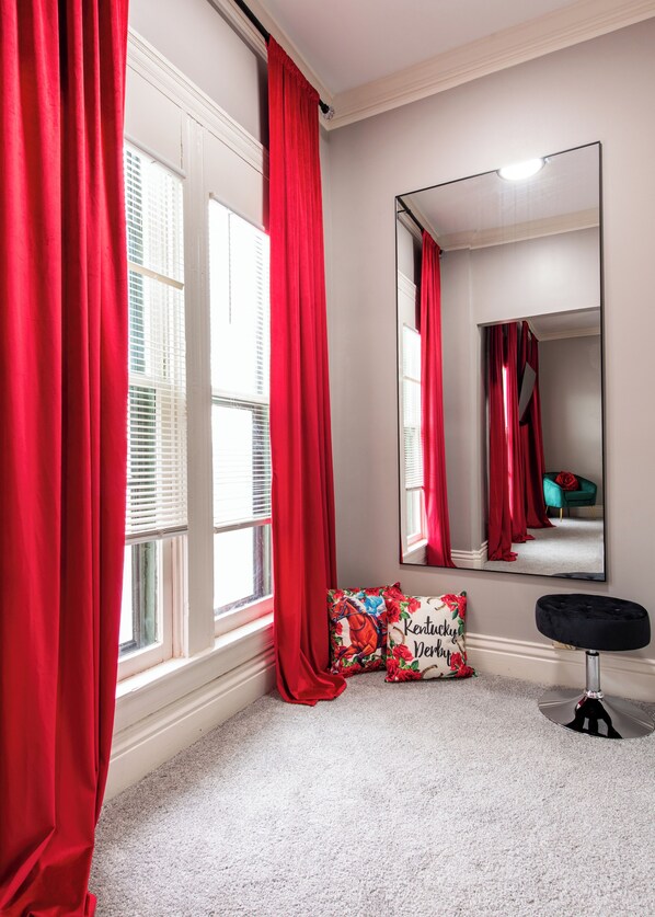 A room with a large mirror, red curtains, a floral pillow, and a black ottoman.