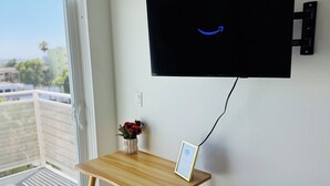Quarto com televisão na parede, uma pequena mesa de madeira com um vaso de flores e um porta-retrato.