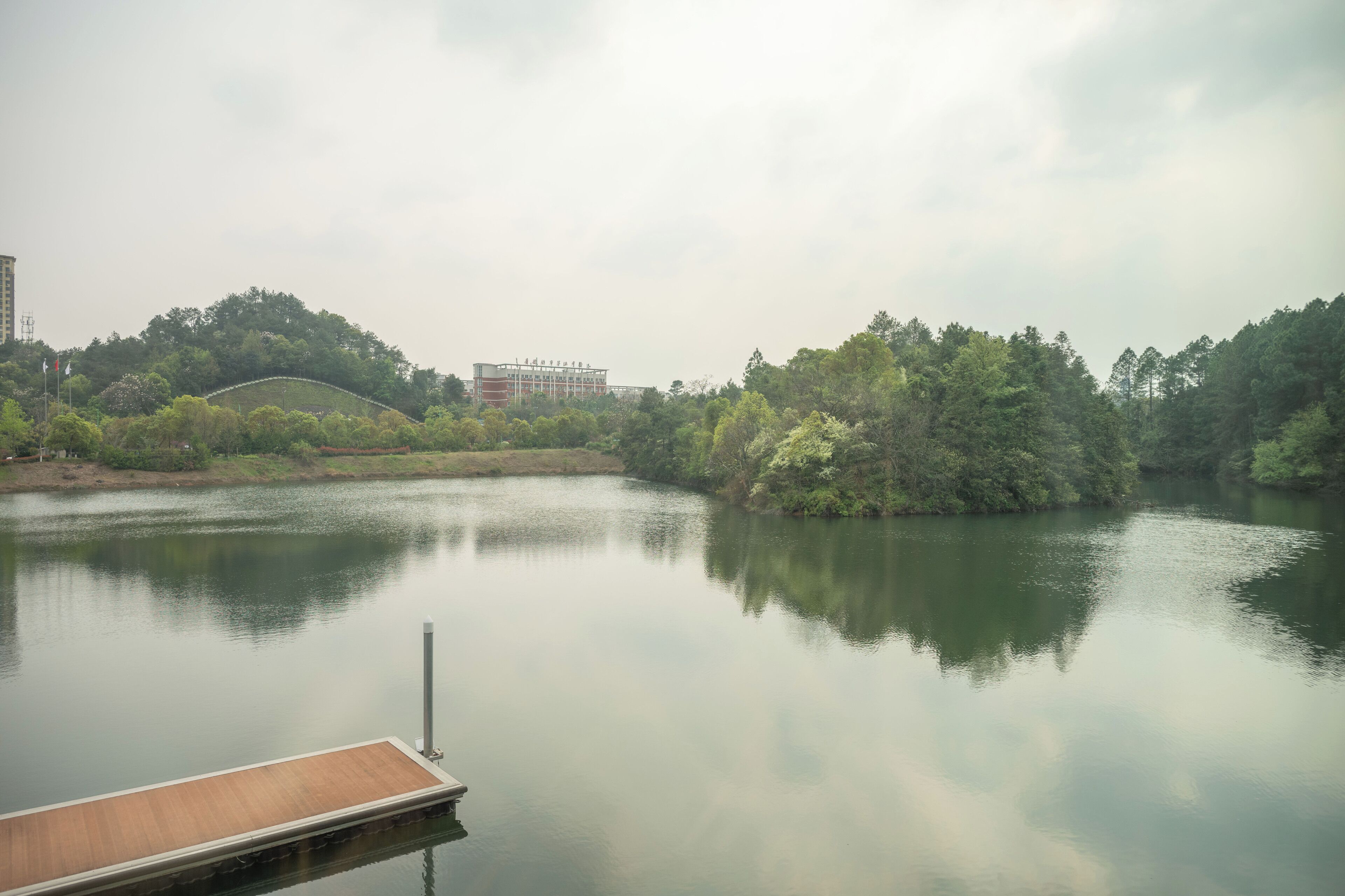 A calm lake with a dock, surrounded by dense greenery and a distant building.