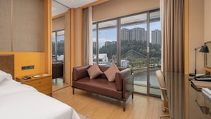 Habitación de hotel con cama, sofá de cuero marrón, escritorio con lámpara y ventana grande que ofrece vistas a edificios y árboles.