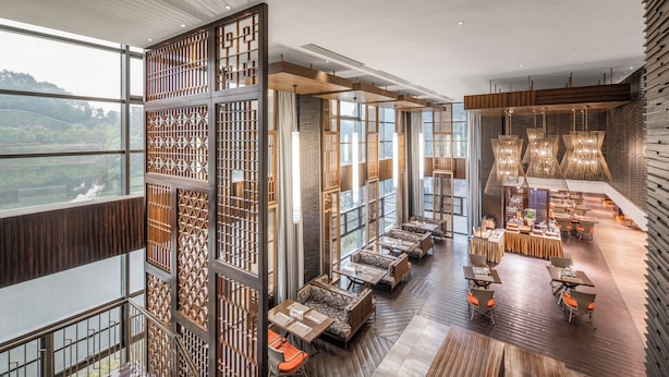 Un interior de hotel moderno con techo alto, ventanas amplias y una zona de comedor central con mesas y sillas.