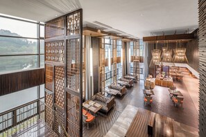 Un interior de hotel moderno con techo alto, ventanas amplias y una zona de comedor central con mesas y sillas.