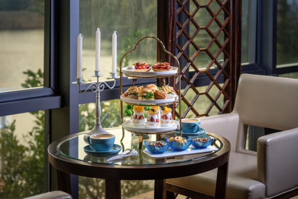 A three-tiered dessert stand with pastries and a candle holder on a glass table, set against a window with a lattice design.