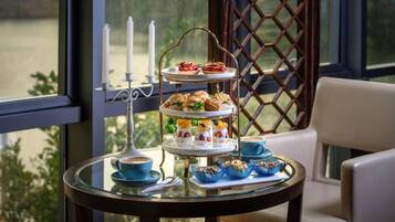 A three-tiered dessert stand with pastries and a candle holder on a glass table, set against a window with a lattice design.