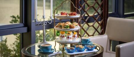 A three-tiered dessert stand with pastries and a candle holder on a glass table, set against a window with a lattice design.