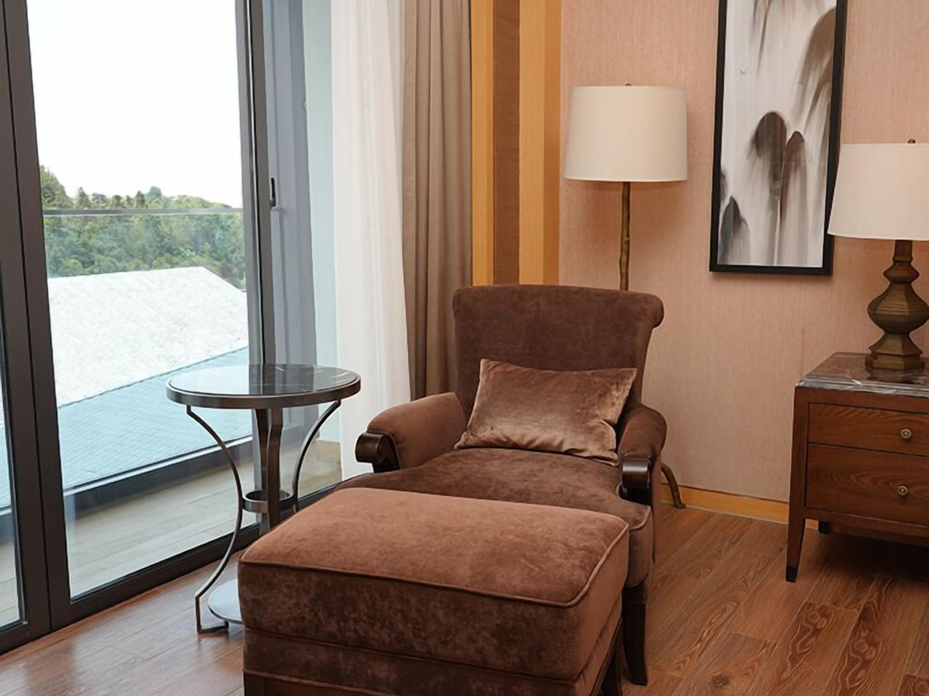 A room with a brown armchair, a small round table, a wooden nightstand, and a large window with a view of greenery.
