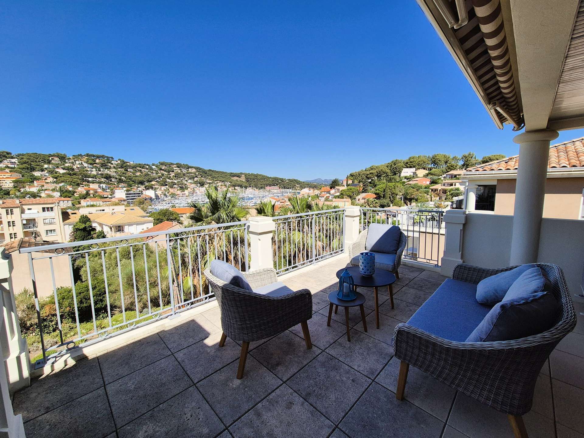 A balcony with wicker furniture, a view of a coastal town, and a clear blue sky.