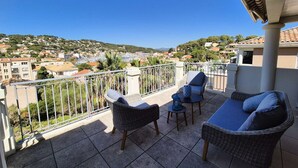 A balcony with wicker furniture, a view of a coastal town, and a clear blue sky.