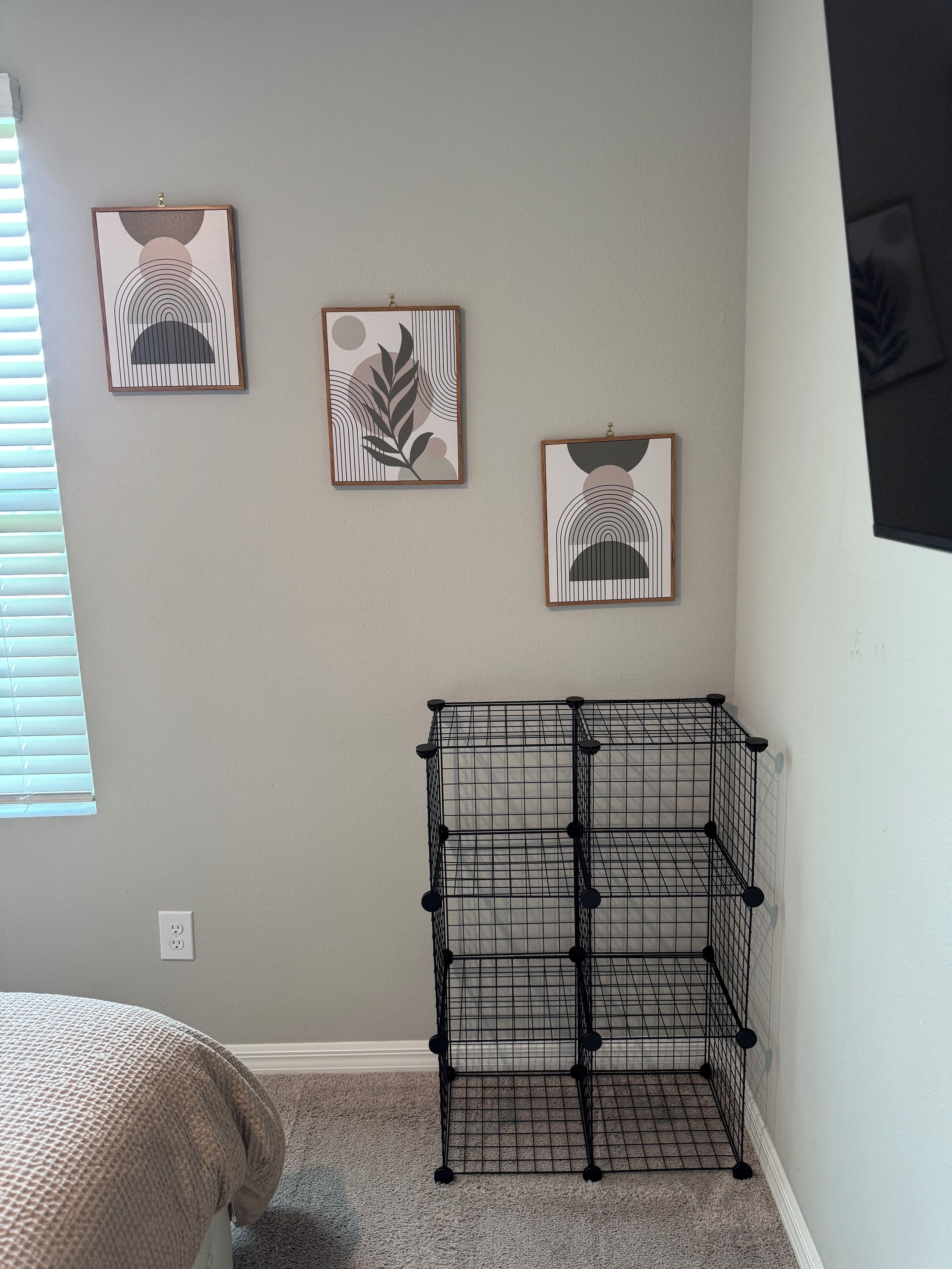 A room with a bed, a black wire shelf unit, and three framed abstract artworks on the wall.