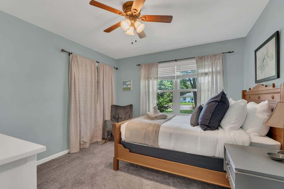 Un dormitorio con cama, escritorio, silla, ventilador de techo y ventana con cortinas.