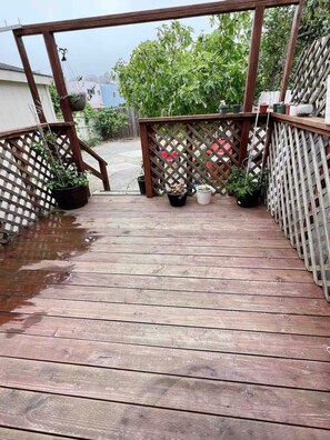 Una terraza de madera con barandilla de celosía, plantas en macetas y vista a una zona residencial.