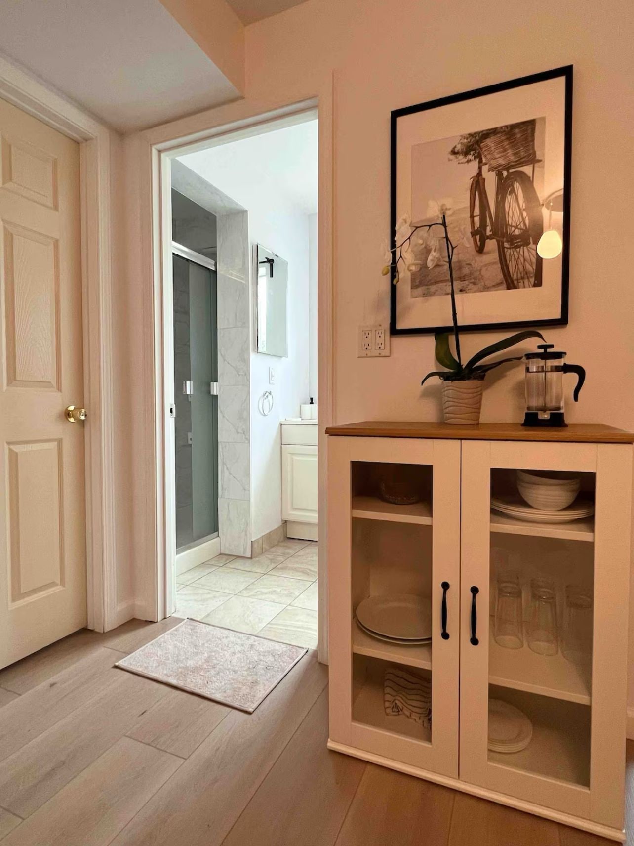 Un pasillo con piso de madera que conduce a un baño con puerta de vidrio para la ducha. Hay un armario de vidrio con platos y vasos.