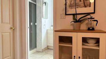Un pasillo con piso de madera que conduce a un baño con puerta de vidrio para la ducha. Hay un armario de vidrio con platos y vasos.
