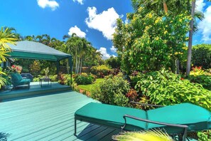 A covered outdoor area with a wooden deck, green lounge chairs, and a variety of tropical plants and trees.