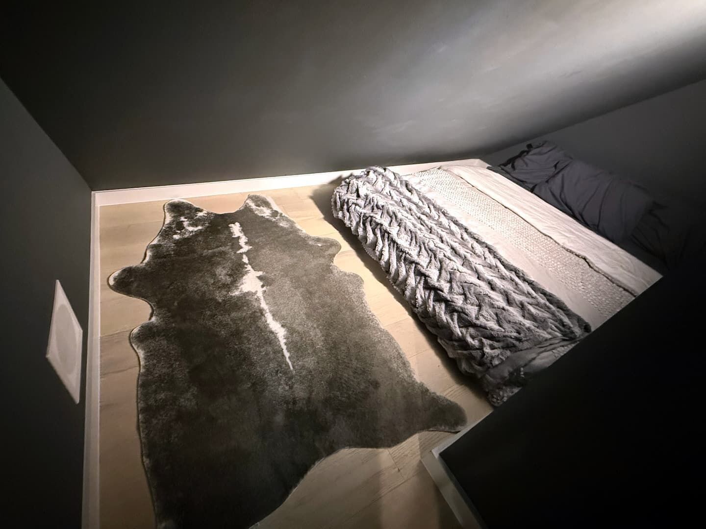 A room with a large rug featuring a Native American design, a bed with a grey blanket, and a wooden floor.