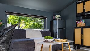 Ett modernt vardagsrum med en grå soffa, ett runt, vitt soffbord och en svart stol. Det finns ett fönster med utsikt över träd, ett svart kylskåp och ett svart skåp med en flätad lucka.