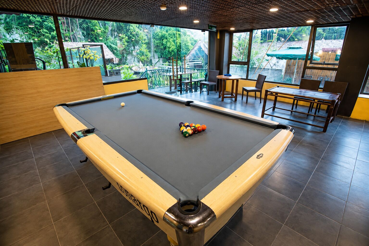 A pool table with a wooden border and a pool cue rack, surrounded by tables and chairs in a room with large windows.