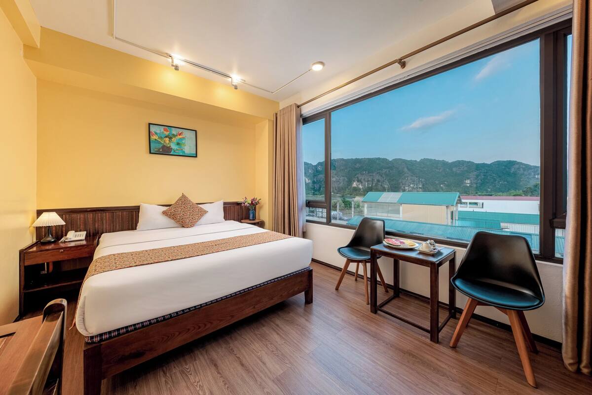 A hotel room with a large bed, a small table, two chairs, a nightstand, and a window with a view of hills.