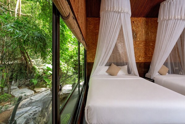 Habitación con dos camas, cada una con ropa blanca y dosel. El cuarto tiene techo y paredes de madera, y un amplio ventanal con vistas a la vegetación.