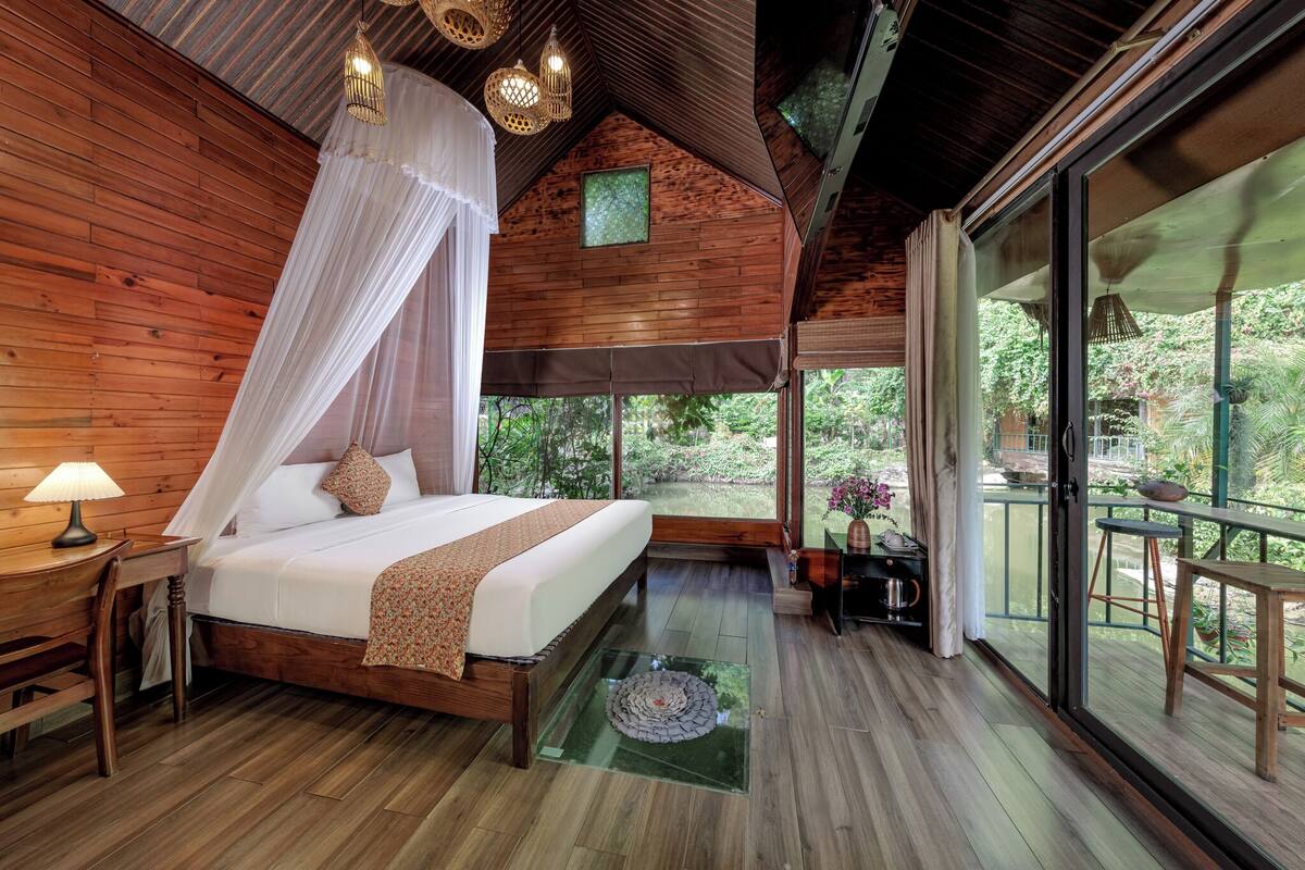 A bedroom with a wooden ceiling, a bed with a canopy, a nightstand, a lamp, and a view of the outdoors through large windows.