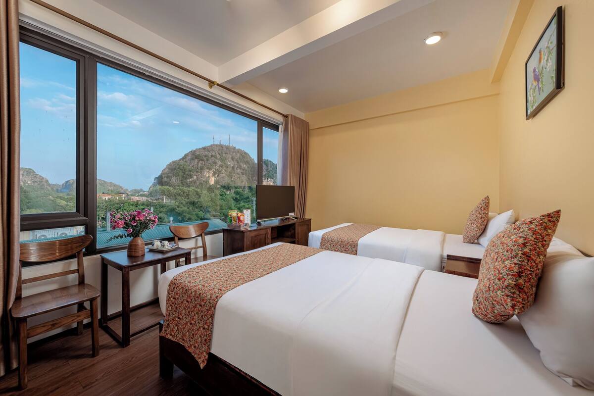 A hotel room with two beds, a desk with a chair, a TV, and a large window offering a mountain view.