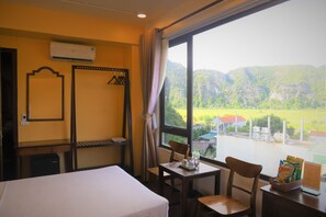 A hotel room with a bed, a table with chairs, a window with a view of mountains, and an air conditioning unit. - Tam Coc Emerald Retreat (Nam Hoa Lu)