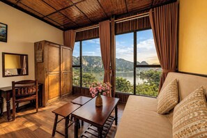A room with a wooden table, a sofa, and a view of a lake and mountains. - Emerald Retreat Tam Coc (Nam Hoa Lu)