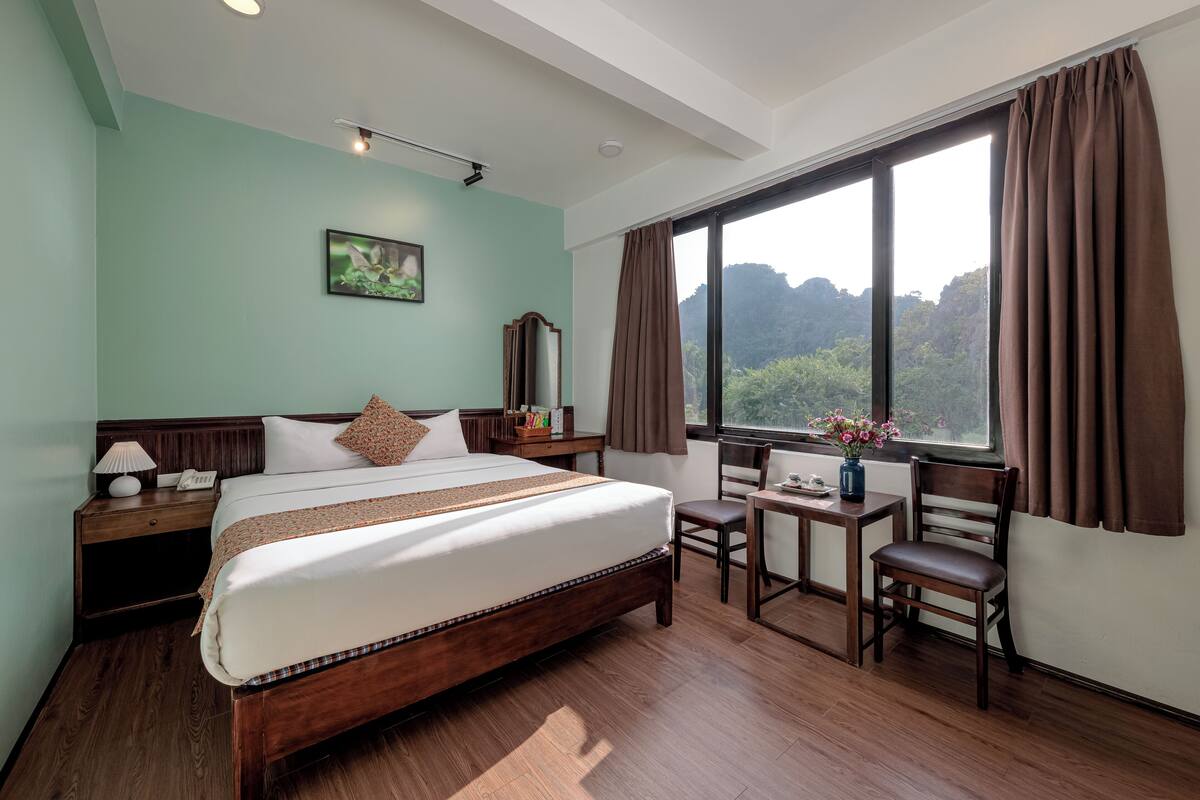 A hotel room with a large bed, a desk, a chair, a nightstand, a lamp, a phone, a mirror, and a window with curtains.
