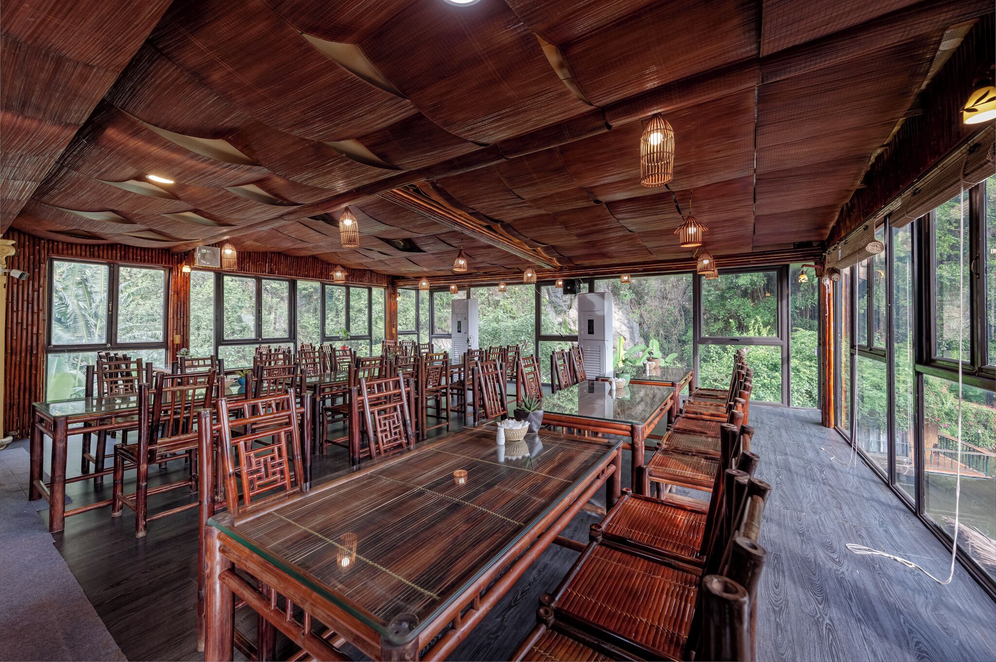 Un comedor espacioso con una larga mesa de madera y sillas, ventanas amplias que ofrecen vistas al bosque y un techo de madera con luces colgantes.