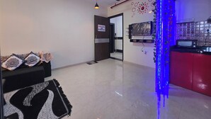 A modern living room with a sofa, a coffee table, and a TV mounted on the wall. There is a kitchen area with a red cabinet and a blue light strip.