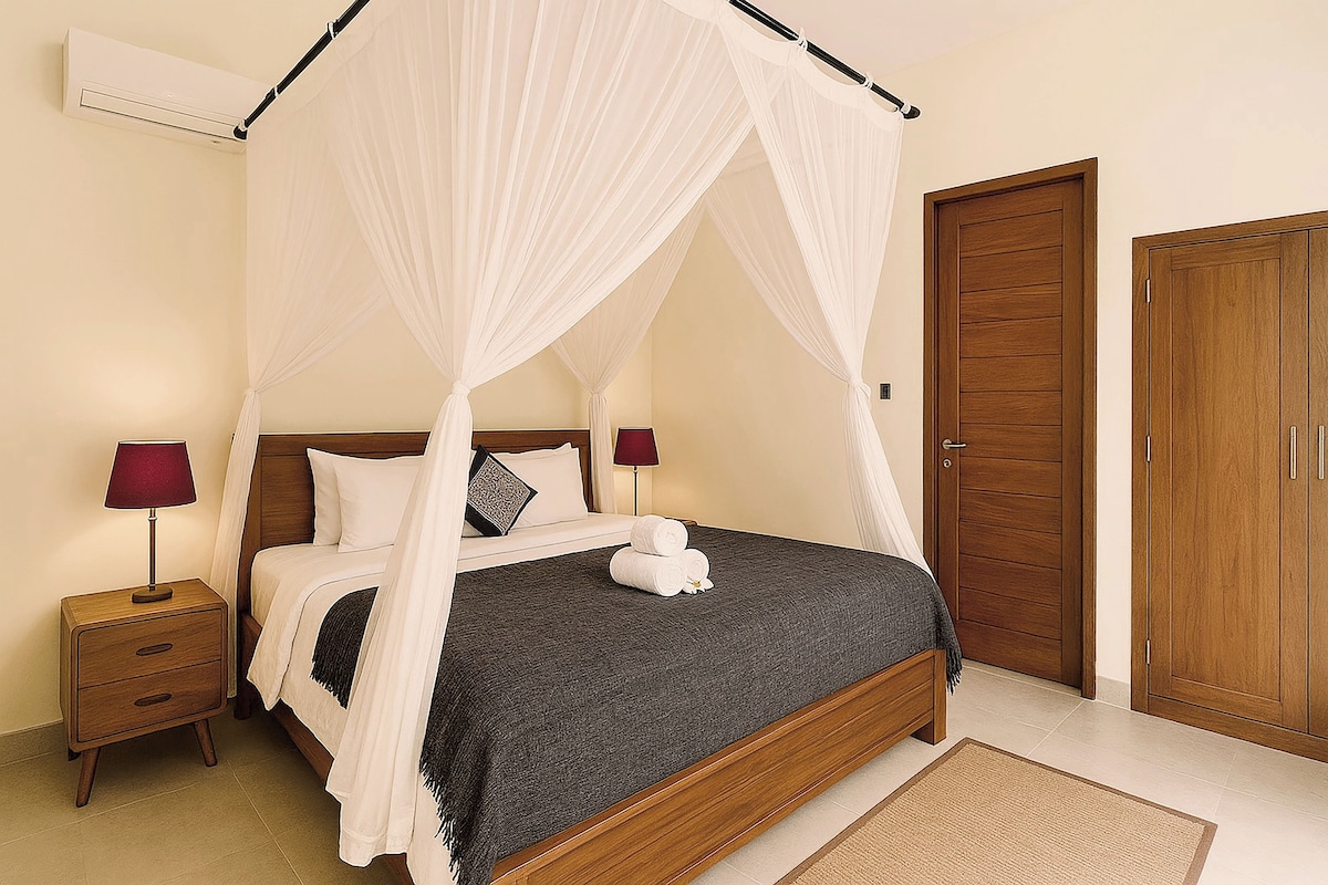 A neatly made bed with a canopy, a wooden nightstand with a lamp, and a closed wooden door.