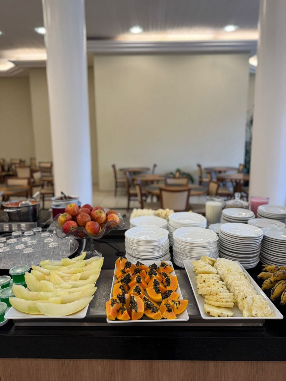 A buffet with various fruits and dishes, including apples, papaya, and pineapple, with plates and bowls stacked in the background.