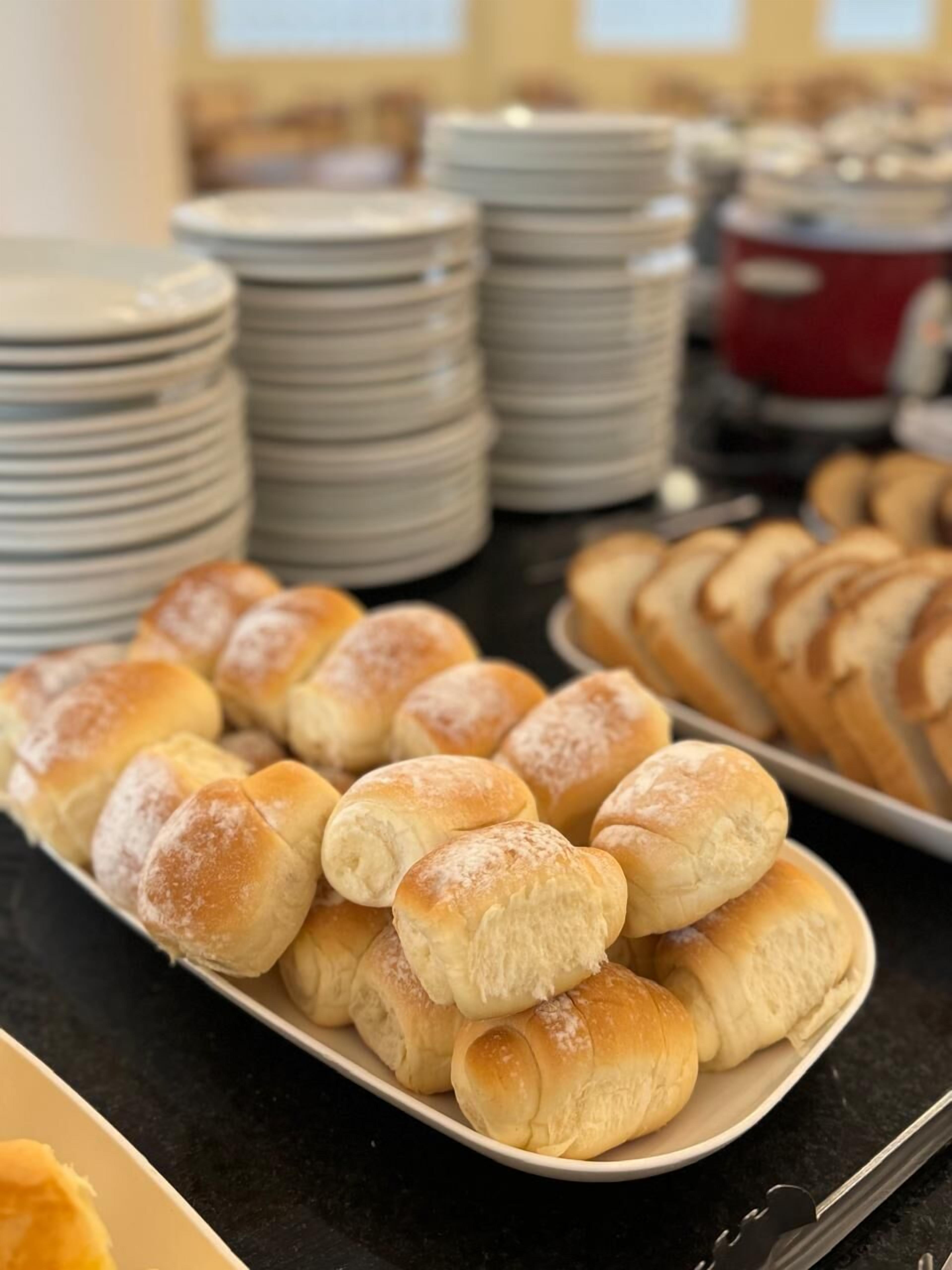 Un tavolo per buffet con diversi prodotti da forno, tra cui panini e dolci, e pile di piatti sullo sfondo.