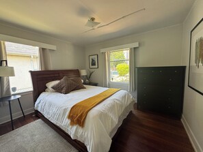 Un dormitorio con cama de madera, una mesita de noche, una lámpara, una ventana con vista a follaje, y un cabecero oscuro.