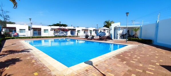 Une piscine rectangulaire, bordée d’une terrasse carrelée, est entourée d’un bâtiment blanc doté de nombreuses fenêtres et d’un espace de détente couvert, aménagé avec des chaises et des tables.