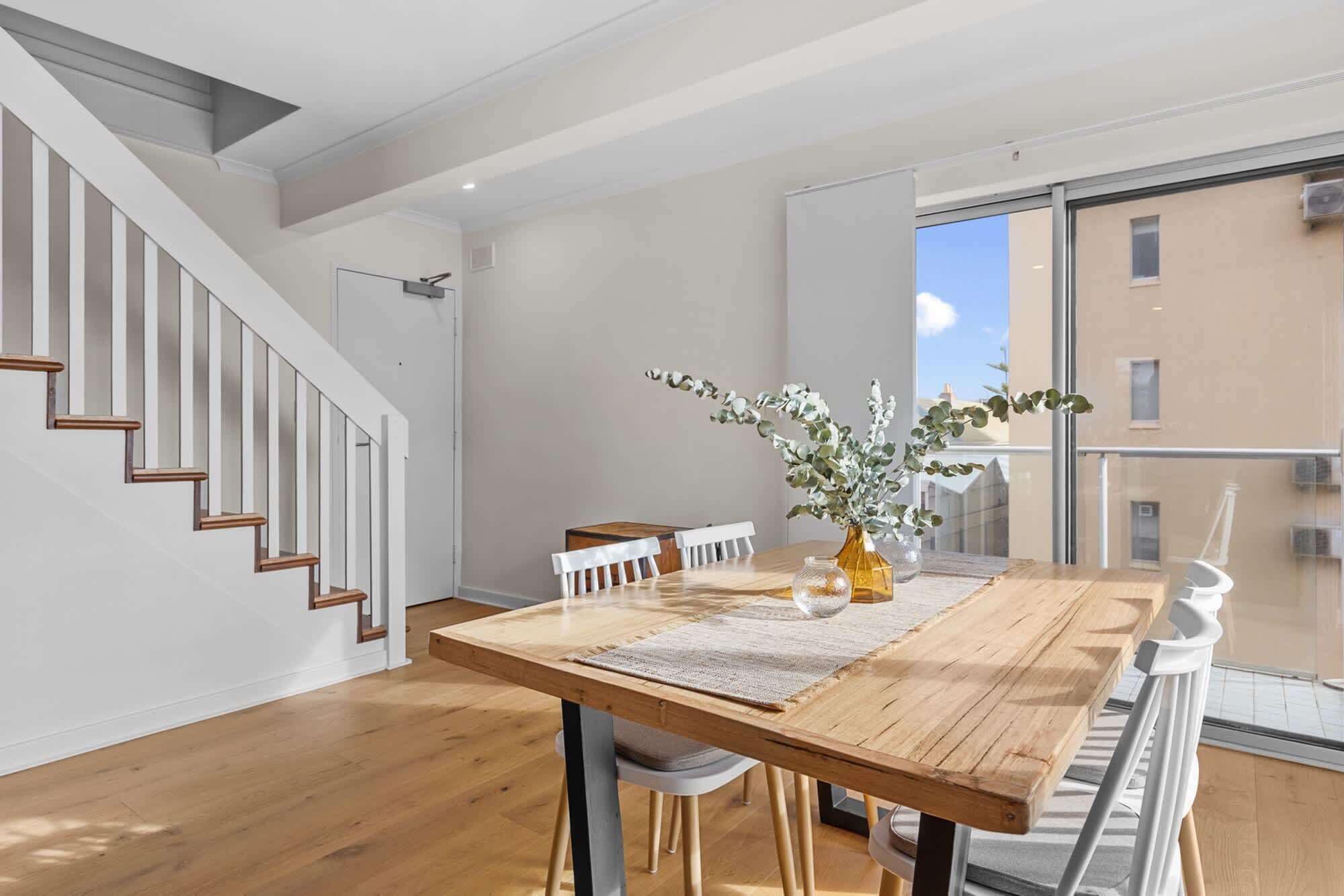 A modern dining area with a wooden table, white chairs, and a vase with greenery. A staircase with wooden steps is visible to the left.
