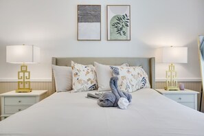 Un lit bien fait, agrémenté de coussins à motifs floraux, d’un ours en peluche gris et de deux tables de chevet avec des lampes.